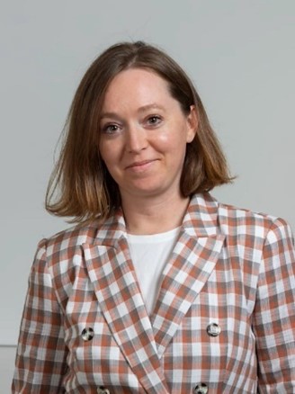 A headshot of Kristen Pudlow. She is in her forties with a plaid orange and white blazer. Her brown hair is cut at shoulder length and she wears a slight grin on her face.